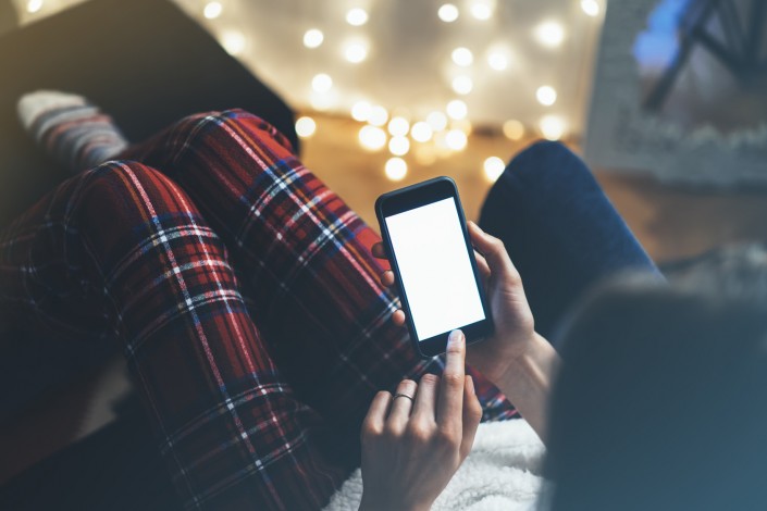 Hipster girl using mobile phone in a home atmosphere, person holding smartphone on background glow bokeh Christmas illumination, hands texting on relax glitter xmas decoration, mockup templates