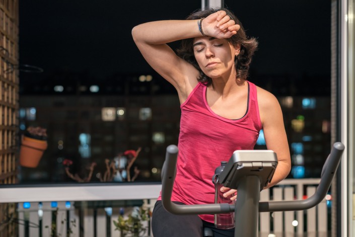 Healthy fit sporty woman exhausted and dizzy rising forearm to forehead at home on exercise static bike after daily female workout routine. Sporty tired girl health weekly beneficial habits.