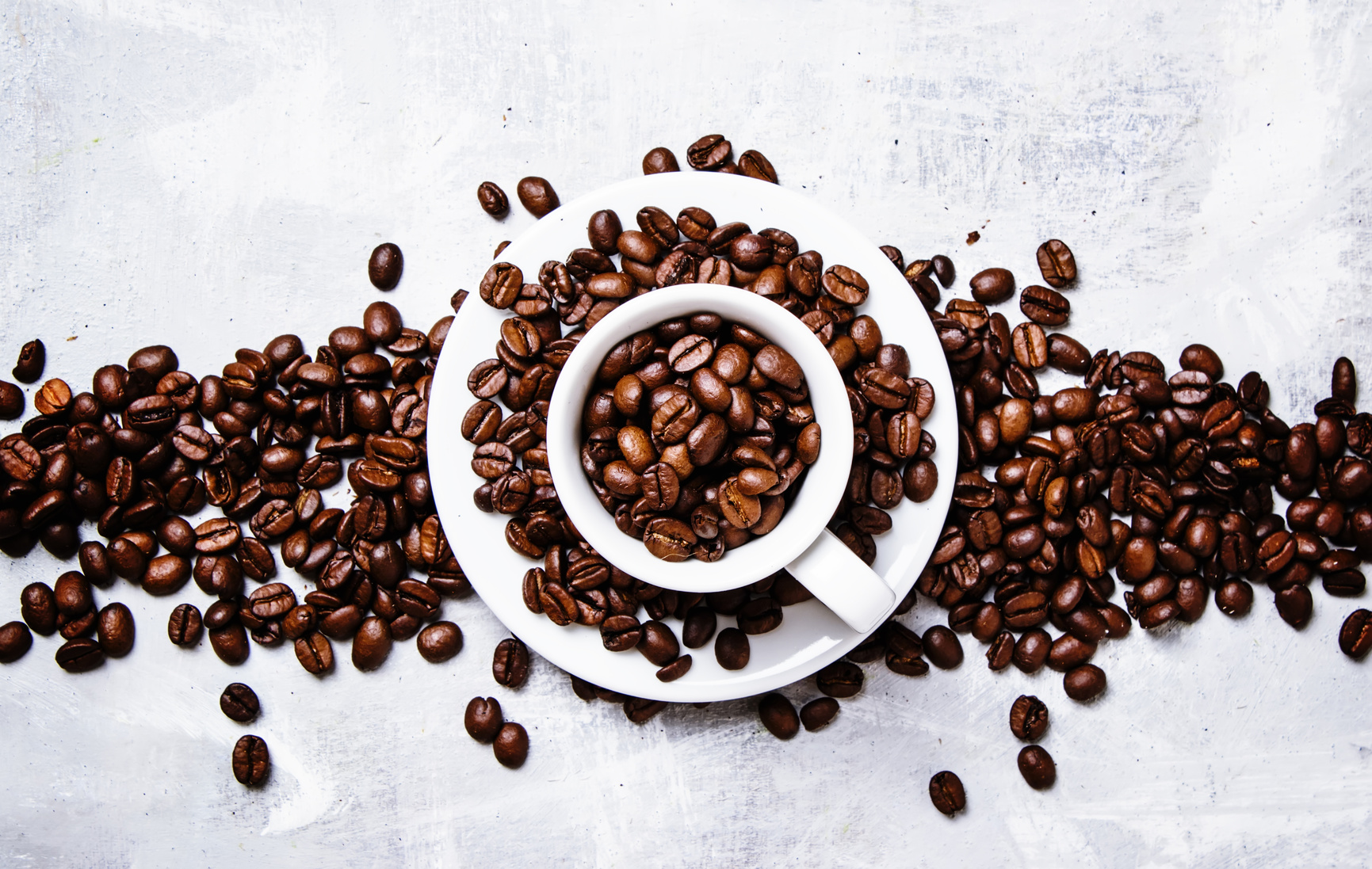 Roasted coffee beans in a white cup and saucer, gray food background, top view, flat lay