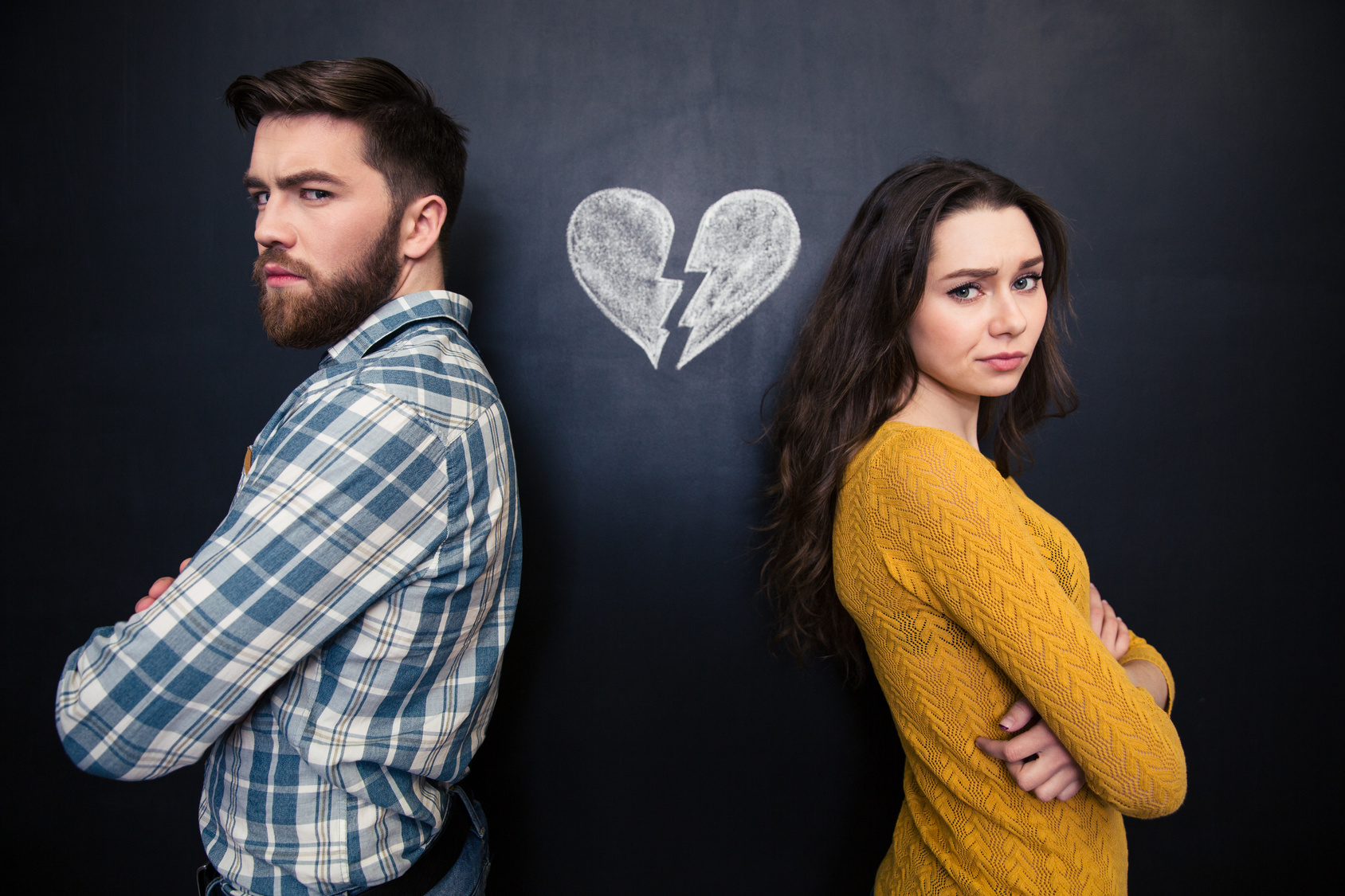 Unhappy couple standing over chalkboard background with drawn broken heart