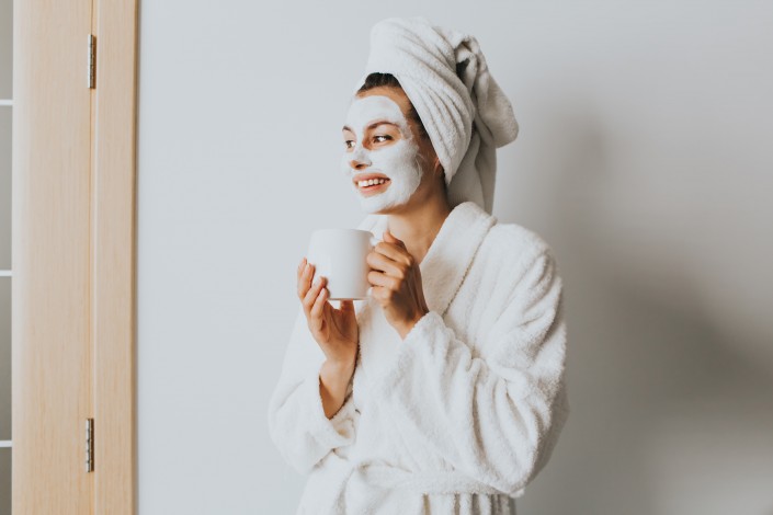 Young woman enjoying coffee at home.Morning tea is my daily routine.Soft photo of fresh young woman in white tender bathrobe and facial mask.Cheerful young woman smiling and looking at the window