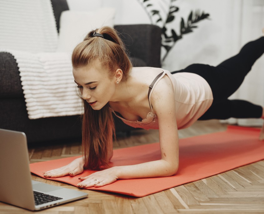 Beautiful young woman with red hair and freckles doing workout exercises at home watching videos on her laptop.