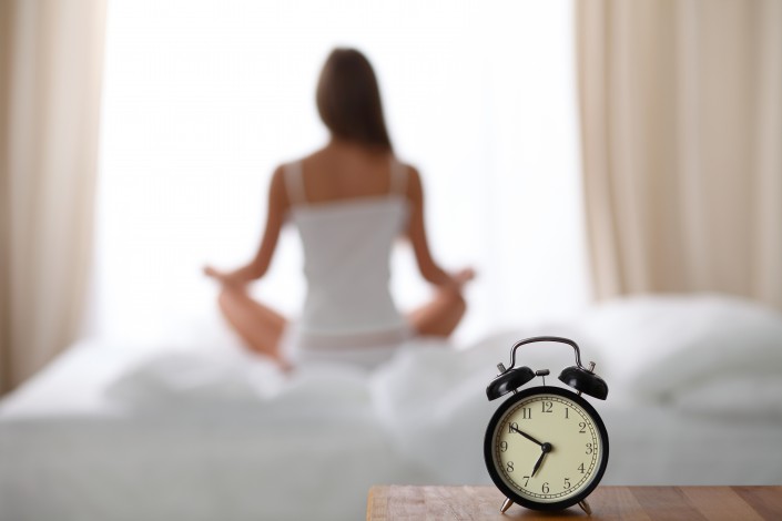 Alarm clock standing on bedside table has already rung early morning to wake up. Woman do yoga in bed in background. Early awakening, healthy lifestyle contemplation concept.