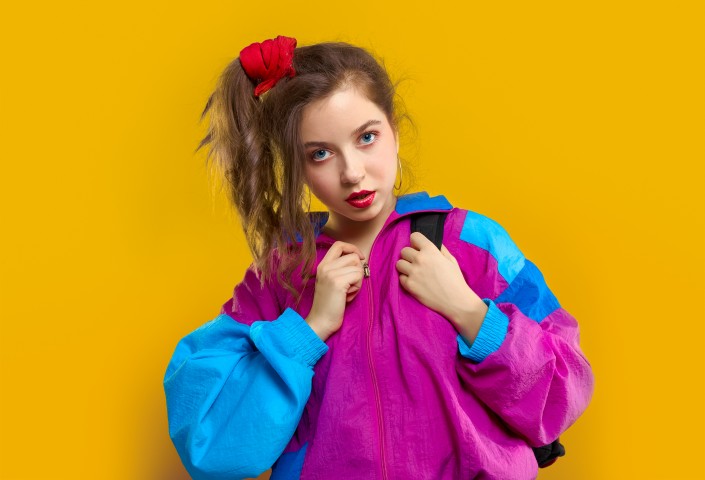 Fashion DJ girl in colorful trendy jacket from 90s. Teenager Girl shows strong face at the disco party of 80s vibes. Fashionable young model on yellow color background.