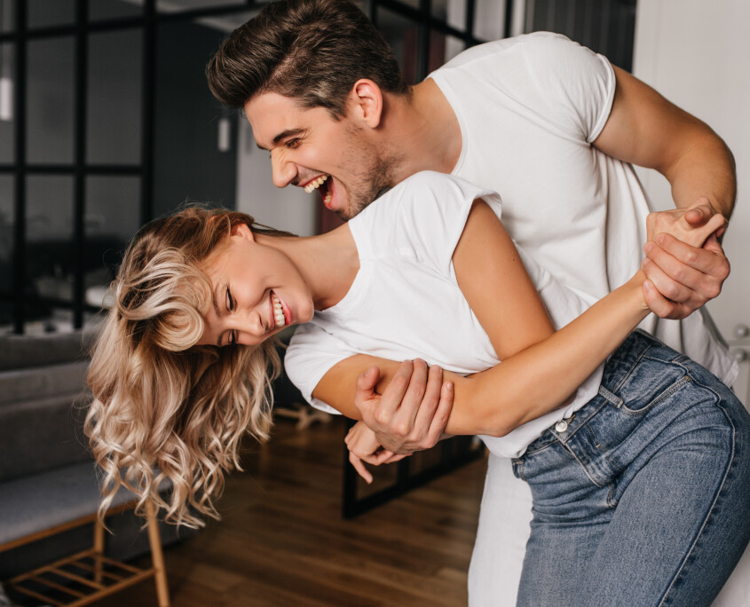 Wonderful girl in white t-shirt dancing with boyfriend. Indoor photo of winsome lady fooling around at home with husband.